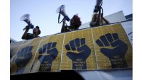Three Black organizers with megaphones above signs that depict a black fist on a yellow background, text on fist says "Not another life"